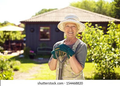 Portrait Of Cheerful Senior Woman With Gardening Tools Outdoors. Older Woman Standing With Shovel In Her Backyard Garden