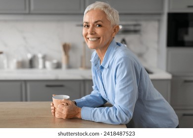 Portrait of cheerful senior woman starting her day with a cup of coffee, finding joy in simple routine of her home kitchen, smiling to camera enjoying hot drink before work - Powered by Shutterstock