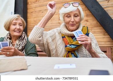 Portrait Of Cheerful Seniors Playing Card Game At Lunch Table On Outdoor Terrace, Focus On Two Old Women Winning Game