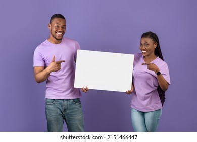Portrait of cheerful smiling black couple holding and pointing fingers at white paper placard for advertisement or text, free copy space for advertising, isolated on violet purple studio background