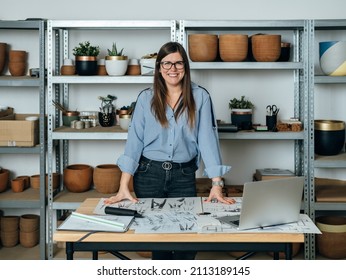 Portrait of Cheerful Smiling Female Furniture Designer Working at her Office. 
Happy business woman standing at her desk with laptop computer, blueprints, sketches and plans and looking at camera.