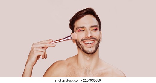 Portrait Of Cheerful Smiling Gay Man With Beard, Pink And Yellow Eyeshadow, No Clothes, Applying Powder On Face. Beauty, Make-up And Skin Care Concept. Studio Shot, Copy Space.