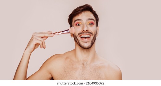 Portrait Of Cheerful Smiling Gay Man With Beard Doing Funny Face, Pink And Yellow Eyeshadow, No Clothes, Applying Powder On Face. Beauty, Make-up And Skin Care Concept. Studio Shot, Copy Space.