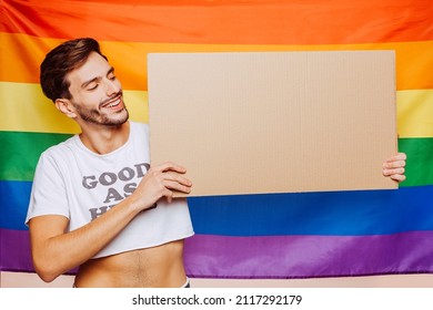 Portrait Of Cheerful Smiling Homosexual Man Holding Empty Blank Board, With A Gay Pride Flag Behind Him At Studio. Lgbtq Flag, Rainbow Flag, Celebrating Parade.