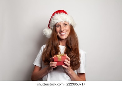 Portrait Of Cheerful Smiling Mature Woman In Christmas Hat Holding Red Gift Box With Gold Ribbons Against White Studio Wall Background