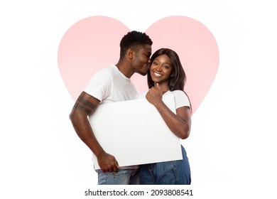 Portrait of cheerful smiling millennial black couple holding white paper placard for advertisement or text, guy kissing lady isolated on white studio background with pink heart shape, free copy space