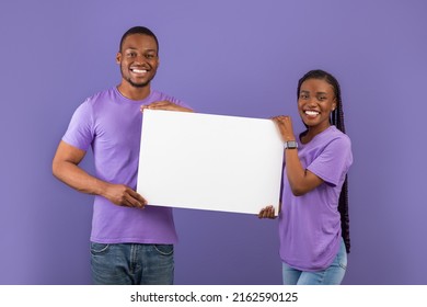 Portrait of cheerful smiling millennial black couple holding white paper placard for advertisement or text together, free copy space for advertising, isolated on violet purple studio background wall