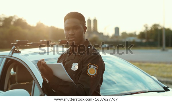Portrait of cheerful smiling policeman holding\
papers leaning on the police car posing at sunlight. happy american\
cop enjoying his work\
outdoors.