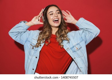 Portrait of a cheerful stylish young woman wearing denim jacket standing isolated over red background, listening to music with headphones, dancing - Shutterstock ID 1408745507