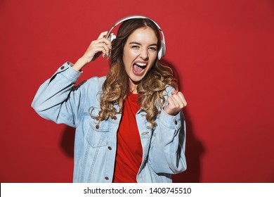 Portrait of a cheerful stylish young woman wearing denim jacket standing isolated over red background, listening to music with headphones, dancing - Shutterstock ID 1408745510