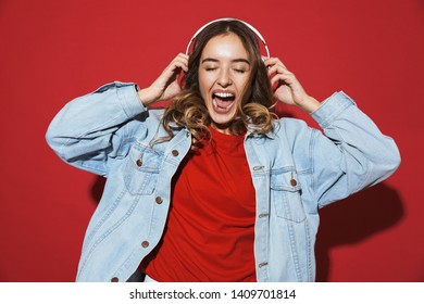 Portrait of a cheerful stylish young woman wearing denim jacket standing isolated over red background, listening to music with headphones, dancing - Shutterstock ID 1409701814