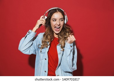 Portrait of a cheerful stylish young woman wearing denim jacket standing isolated over red background, listening to music with headphones, dancing - Shutterstock ID 1409701817