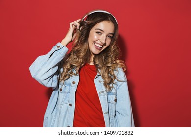 Portrait of a cheerful stylish young woman wearing denim jacket standing isolated over red background, listening to music with headphones - Shutterstock ID 1409701832
