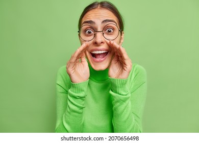 Portrait of cheerful surprised mysterious young woman keeps hands near mouth wshispers secret wears round spectaclesandd casual jumper shares gossips with you isolated over green background.