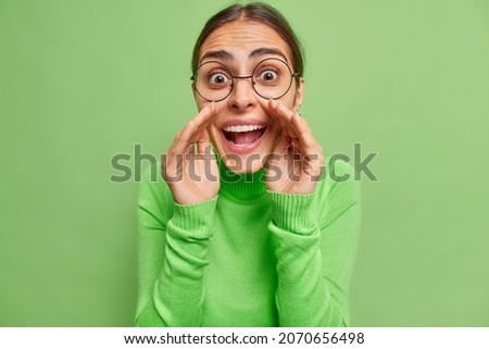 Portrait of cheerful surprised mysterious young woman keeps hands near mouth wshispers secret wears round spectaclesandd casual jumper shares gossips with you isolated over green background. Stock foto © 