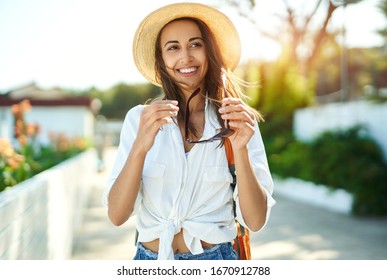 Portrait fröhliche Touristenfrau geht alte Straße. Attraktive Frau in Strohhut, weißes Hemd mit Sonnenbrille, genießt Solourlaub in Europa. Tourismus und Reisekonzept.