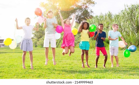 Portrait Of Cheerful Tween Girls And Boys Holding Colorful Balloons In Hands Jumping Together In Summer City Park. Concept Of Happy Preadolescence