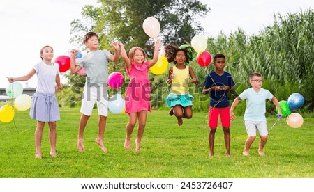 Portrait of cheerful tween girls and boys holding colorful balloons in hands jumping together in summer city park. Concept of happy preadolescence