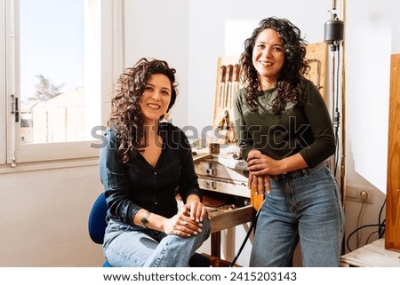 Portrait of cheerful twin sisters with curly hair working as jewelers in creative workshop in sunlight looking at camera