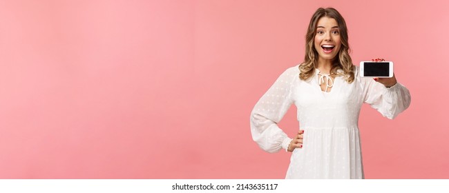 Portrait Of Cheerful, Upbeat Attractive Blond Caucasian Girl In White Dress, Showing Smartphone Display, Hold Mobile Phone Horizontal Smiling Camera Amazed, Stand Pink Background