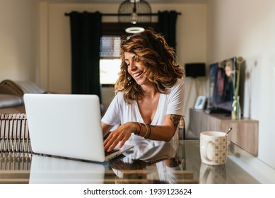 Portrait Of A Cheerful Woman At Home With A Laptop And A Cup Of Coffee On The Table. 30 Year Old Girl Connected Online To A Meeting With Friends And Family. Telework Concept