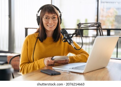 Portrait of cheerful woman host streaming audio podcast using microphone and laptop in broadcast studio, holding notepad, looking at camera and smiling. Young female podcaster running her radio show