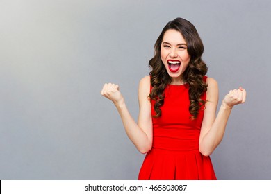 Portrait of a cheerful woman in red dress celebrating her success over gray background