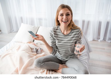 Portrait of cheerful woman is relaxing while baby is sleeping. She is holding glass of water and cellphone and looking at camera with joy - Powered by Shutterstock