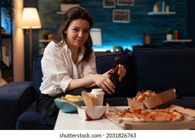 Portrait Of Cheerful Woman Sitting On Couch Smiling At Camera While Enjoying Home-delivered Late At Night In Living Room. Caucasian Female Holding Beer Bottle. Takeaway Food Delivery