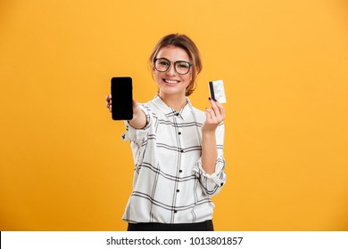 Portrait Of Cheerful Woman Wearing Eyeglasses Holding Smartphone And Credit Card In Hands Isolated Over Yellow Background