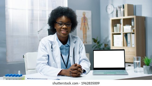 Portrait of cheerful young African American woman physician in white medical coat sitting at table in clinic cabinet showing laptop with blank screen speaking on online video chat, healthcare webinar