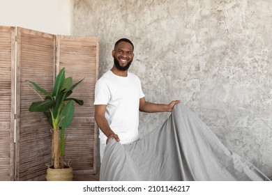 Portrait of cheerful young African American man making bed after wake up feeling rested, happy bearded black guy changing gray linen flax bedsheets at home standing in modern bedroom looking at camera