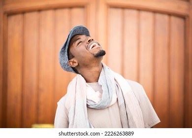 Portrait of cheerful young african man with cap and scarf looking up and laughing - Shutterstock ID 2226263889
