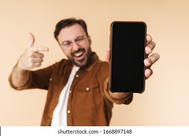 Portrait of a cheerful young arttractive bearded man wearing casual clothes standing isolated over beige background, showing blank screen mobile phone, pointing finger