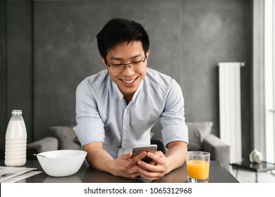 Portrait Of A Cheerful Young Asian Man Using Mobile Phone While Having Breakfast On A Kitchen At Home