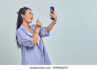 Portrait of cheerful young Asian woman using mobile phone taking selfie and showing heart sign with two fingers crossed isolated on white background