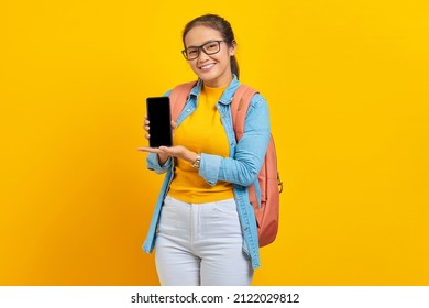 Portrait Of Cheerful Young Asian Woman Student In Casual Clothes With Backpack  Showing Blank Screen Mobile Phone Isolated On Yellow Background. Education In College University Concept