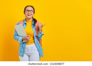 Portrait Of Cheerful Young Asian Woman Student In Casual Clothes With Backpack  Holding Laptop And Pointing Thumbs Up Aside Isolated On Yellow Background. Education In University College Concept