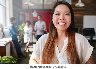 Portrait Of Cheerful Young Asian Woman Standing In Office With Coworkers Talking In Background. Female Creative Professional Looking At Camera And Smiling.