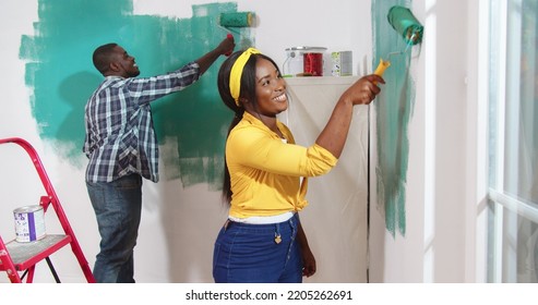 Portrait Of Cheerful Young Beautiful African American Woman Smiling Painting Walls In Green Color With Paint Roller Brush, Helping Her Husband To Redesign Room. Side View, Home Renovation, Repair