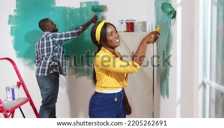 Portrait of cheerful young beautiful African American woman smiling painting walls in green color with paint roller brush, helping her husband to redesign room. Side view, home renovation, repair