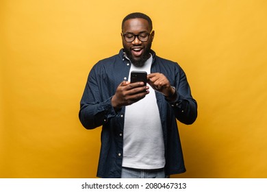 Portrait of cheerful young black guy sending message on mobile phone against yellow background. Happy African-American man using smartphone, enjoying chatting online, messaging in social networks - Shutterstock ID 2080467313