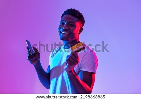 Portrait of cheerful young black man holding cellphone and credit card, using banking or shopping mobile app, receiving cashback, trading or gambling online in neon light