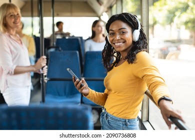 Portrait of cheerful young black woman passenger standing in bus near window wearing wireless headphones using cell moving in modern tram, enjoying trip at public transport posing looking at camera - Powered by Shutterstock