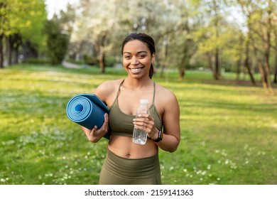 Portrait of cheerful young black woman holding fresh water and sports mat at urban park. Fit African American lady ready for yoga practice, keeping hydrated during outdoor training - Powered by Shutterstock