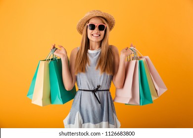 Portrait of a cheerful young blonde woman in summer hat and sunglasses holding shopping bags isolated over yellow background - Powered by Shutterstock