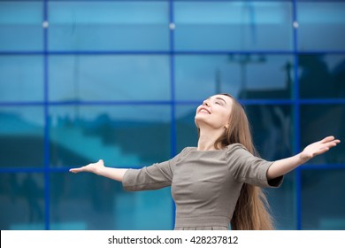 Portrait of cheerful young business woman standing with outstretched arms and relaxing outdoors with closed eyes. Happy beautiful caucasian woman feeling ecstatic on the street in summer. Copy space