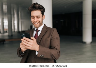 Portrait of cheerful young businessman in suit checking using mobile phone smiling looking screen while standing outside contemporary office building. Concept of business communication and technology. - Powered by Shutterstock