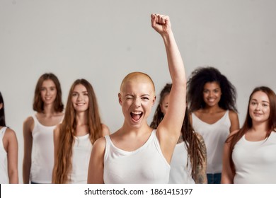 Portrait of cheerful young caucasian woman with shaved head in white shirt raised her arm while winking at camera. Diverse women standing over grey background. Diversity concept. Selective focus