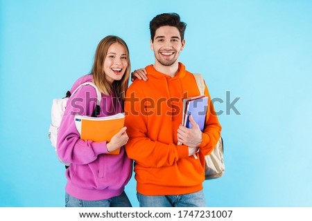 Portrait of a cheerful young couple of students wearing backpacks, carrying textbooks standing isolated over blue background